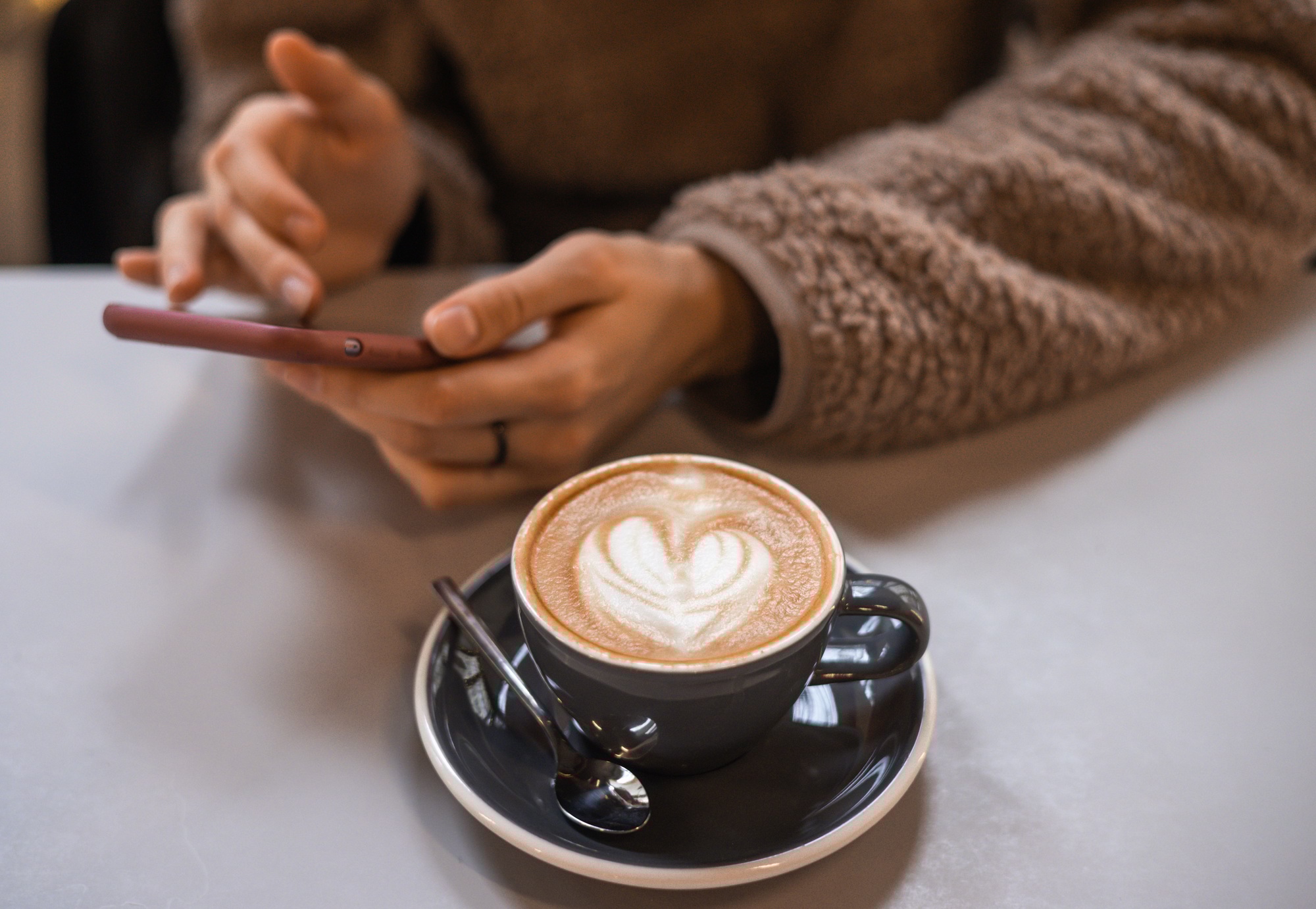 Girl in fluffy sweater holds a smartphone in hands and writes message next to a cup of coffee