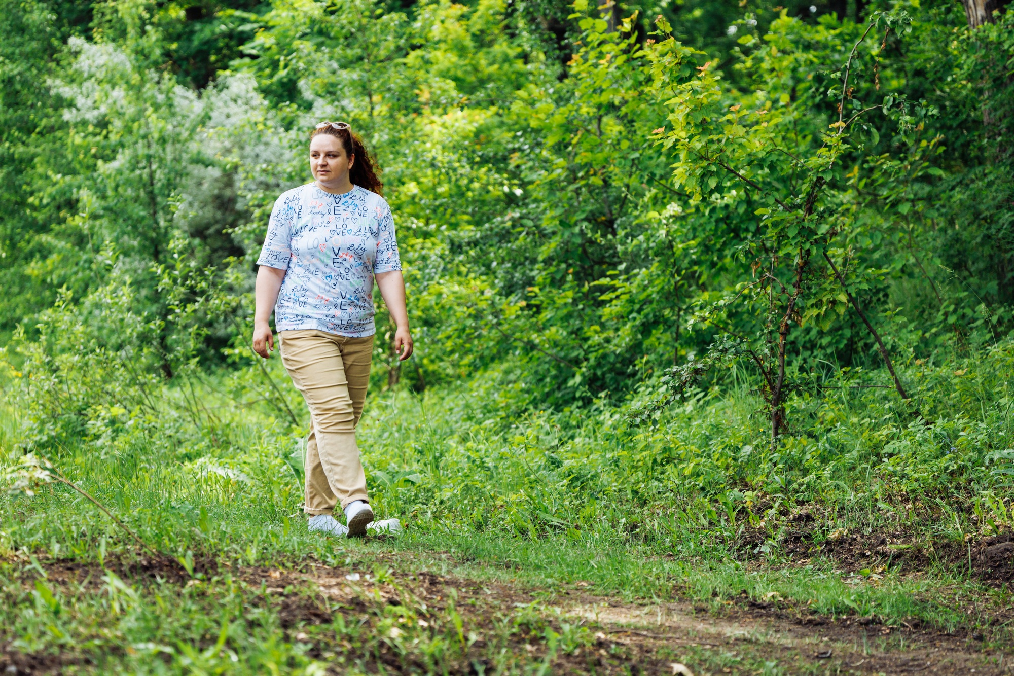Portrait of young fat woman with long curly red hair walking on path in park forest among green